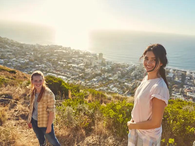 Zwei junge Frauen einer Gruppenreise genießen auf dem Signal Hill den sonnigen Panoramablick über Kapstadt und das Meer.