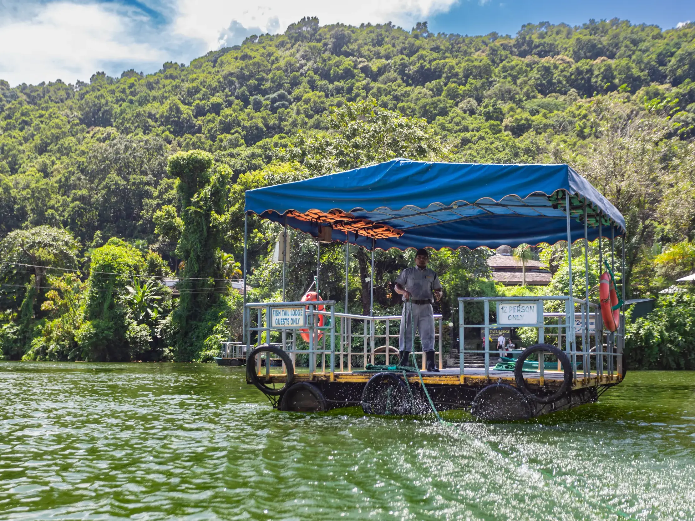 Eine handgezogene Fähre mit blauem Dach gleitet über den Phewa-See in Pokhara zur Fish Tail Lodge vor grünen Hügeln Nepals.