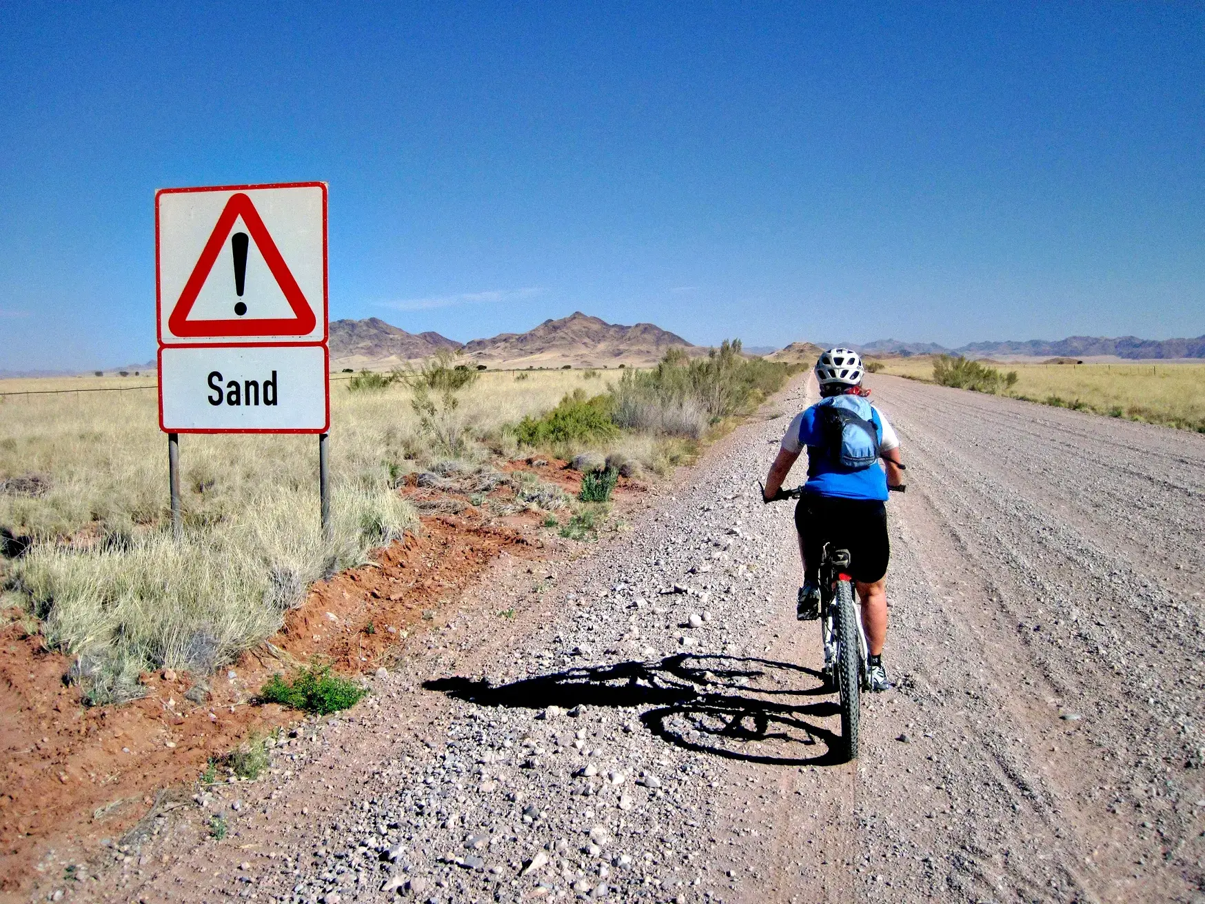 Radfahrerin auf einer Schotterpiste in der Namib-Wüste. Ein Schild warnt vor Sand, im Hintergrund erheben sich Berge.