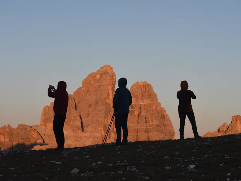 Eine Gruppe Wanderer fotografiert das Alpenglühen der markanten Drei Zinnen während einer Hüttenwanderung in den Dolomiten.