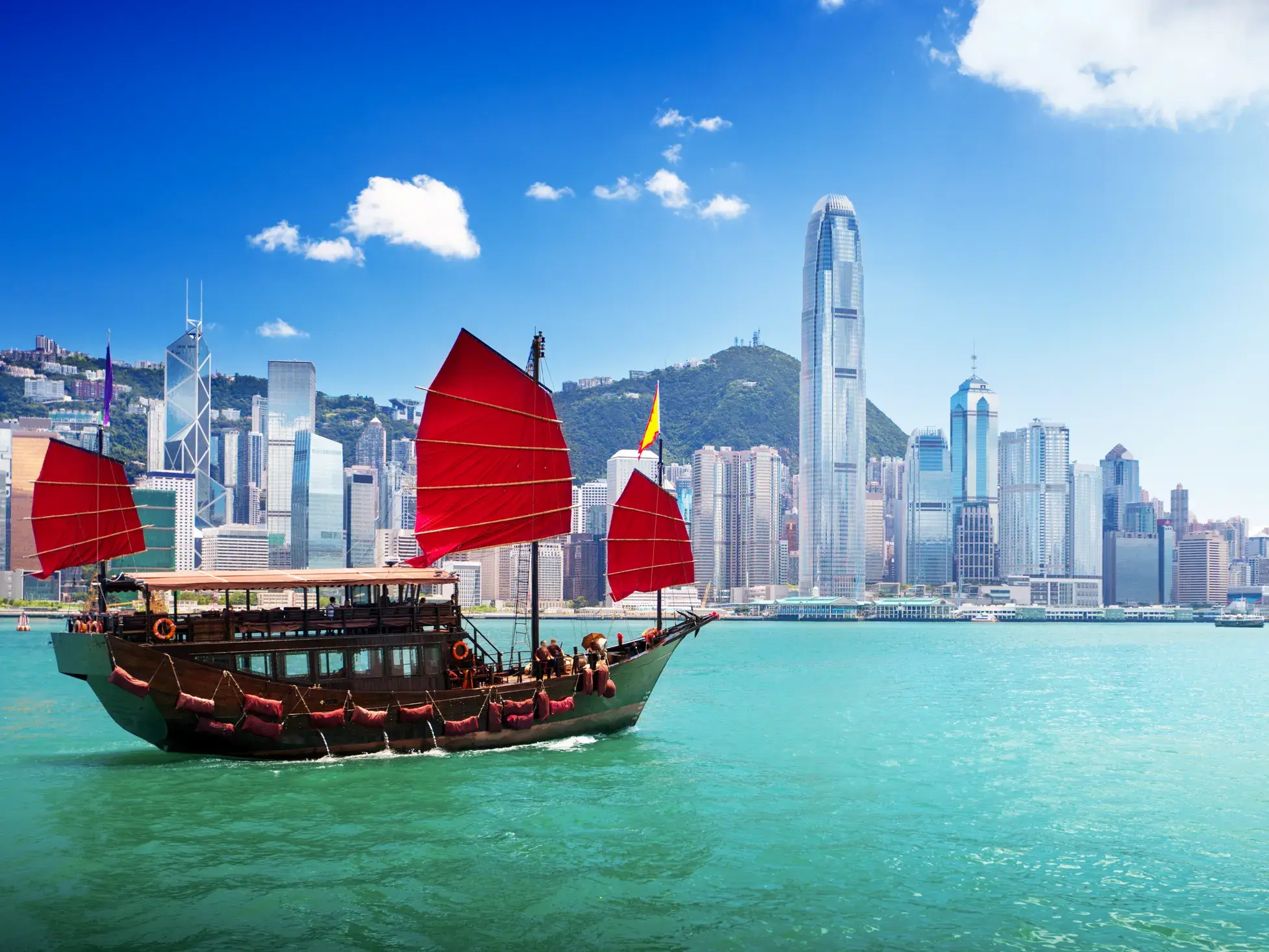 Eine traditionelle chinesische Dschunke mit roten Segeln fährt im Victoria Harbour vor der modernen Skyline von Hongkong.
