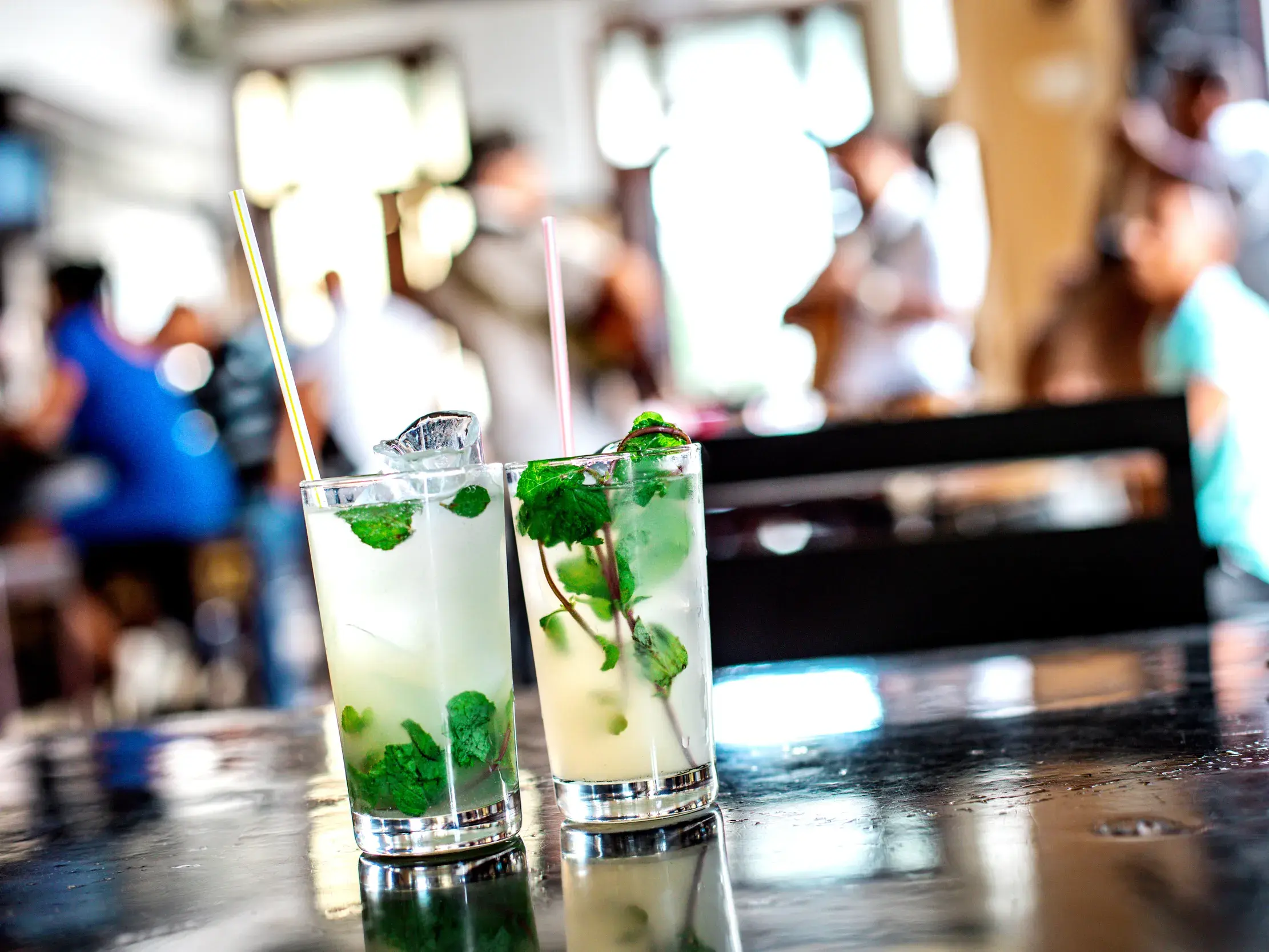 Zwei erfrischende Mojito-Cocktails mit Minze und Eiswürfeln auf einem dunklen Tisch in einer belebten Bar in Havanna.