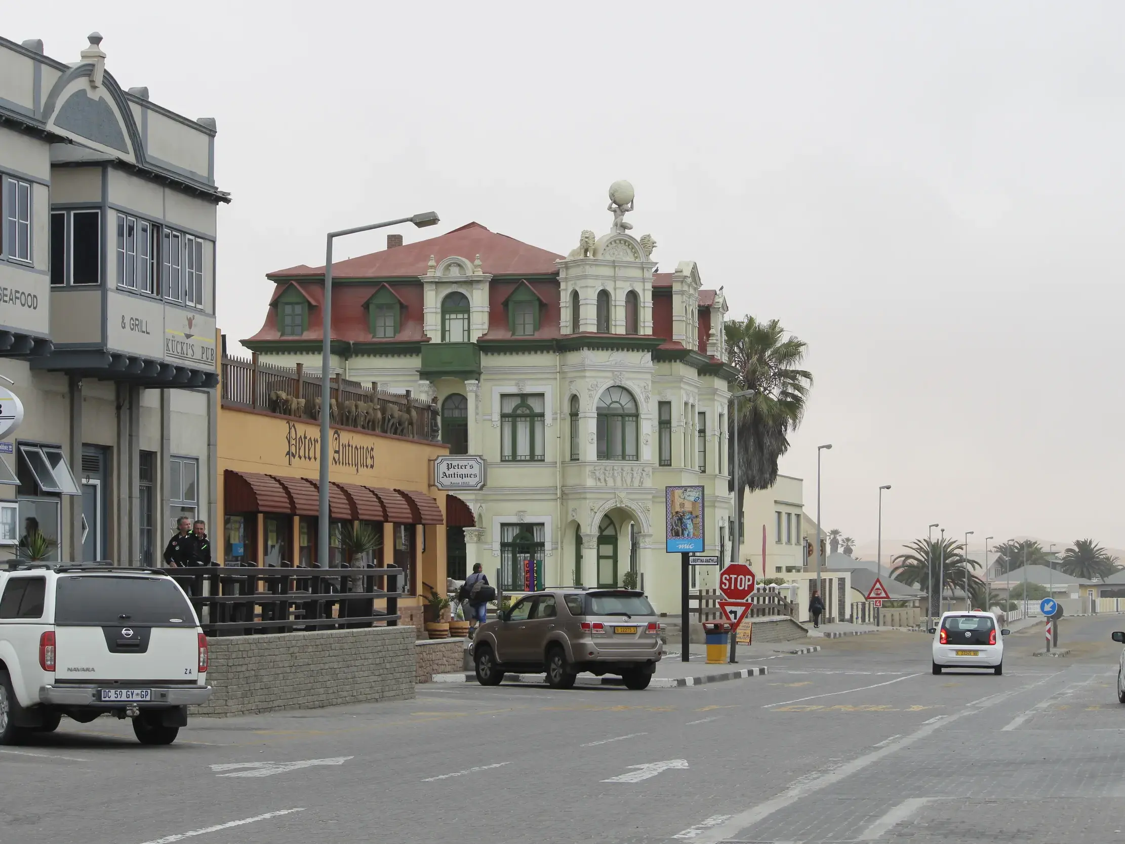 Straßenszene in Swakopmund mit historischem Gebäude im deutschen Kolonialstil, Autos und Passanten am Straßenrand.