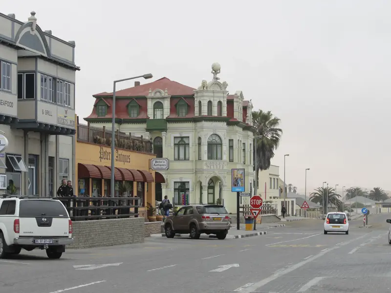 Straßenszene in Swakopmund mit historischem Gebäude im deutschen Kolonialstil, Autos und Passanten am Straßenrand.