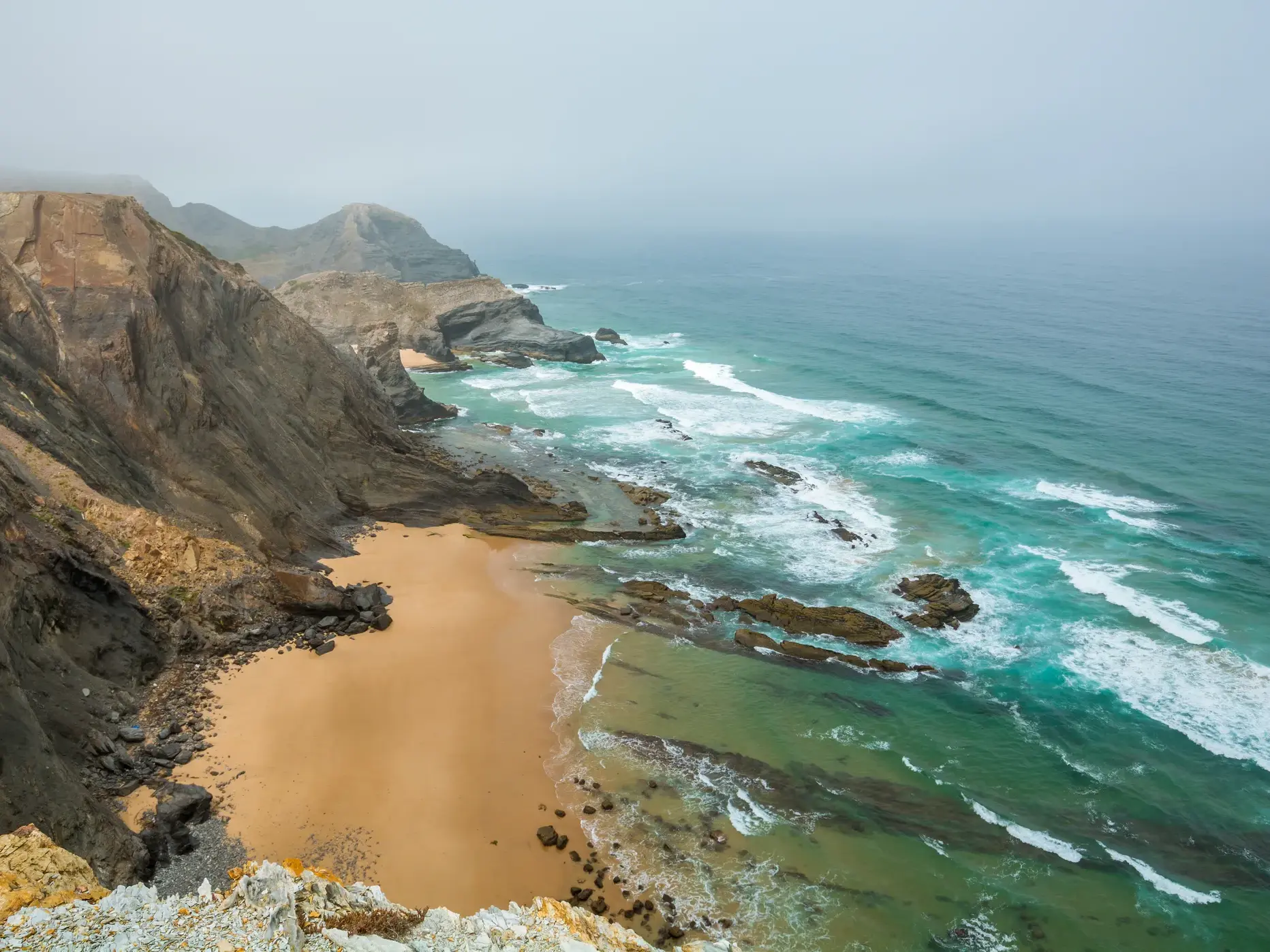 Blick von den Klippen des Fischerpfads auf die raue Atlantikküste der Algarve mit Sandstrand und türkisem Wasser.