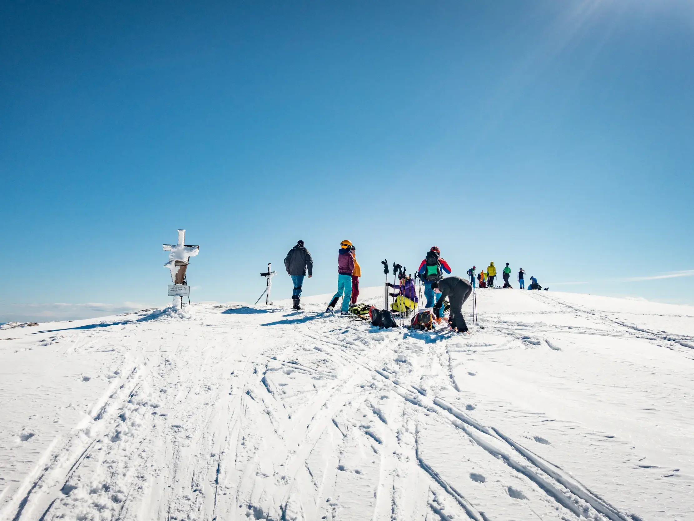 Eine Gruppe Winterwanderer macht bei Sonnenschein eine Rast am Gipfelkreuz des Jenner im Nationalpark Berchtesgaden.