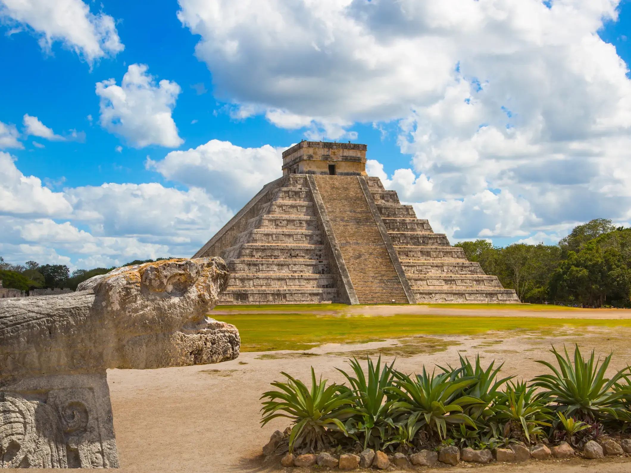 Die Kukulcán-Pyramide in Chichén Itzá an einem sonnigen Tag, im Vordergrund eine steinerne Schlangen-Skulptur.