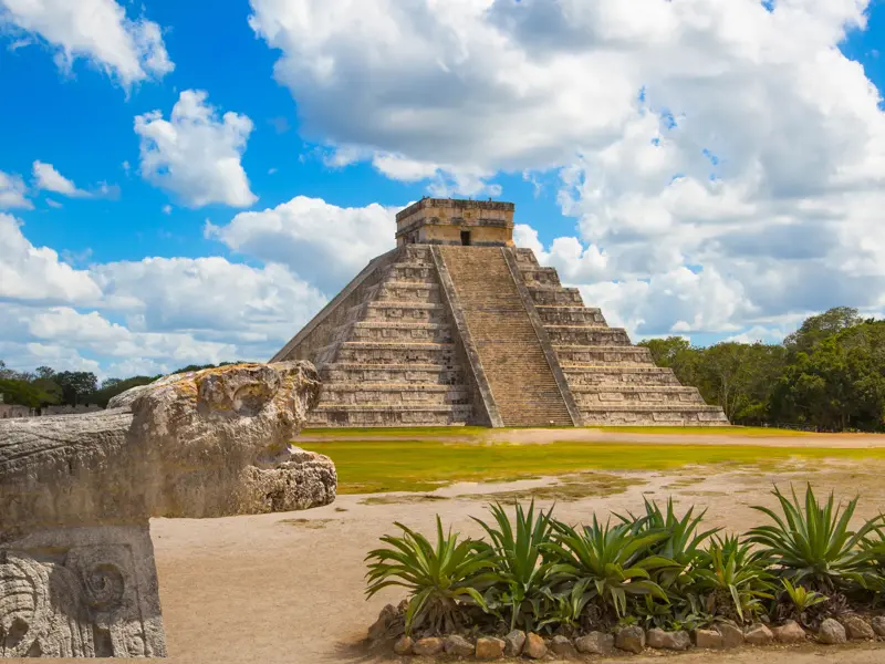 Die Kukulcán-Pyramide in Chichén Itzá an einem sonnigen Tag, im Vordergrund eine steinerne Schlangen-Skulptur.