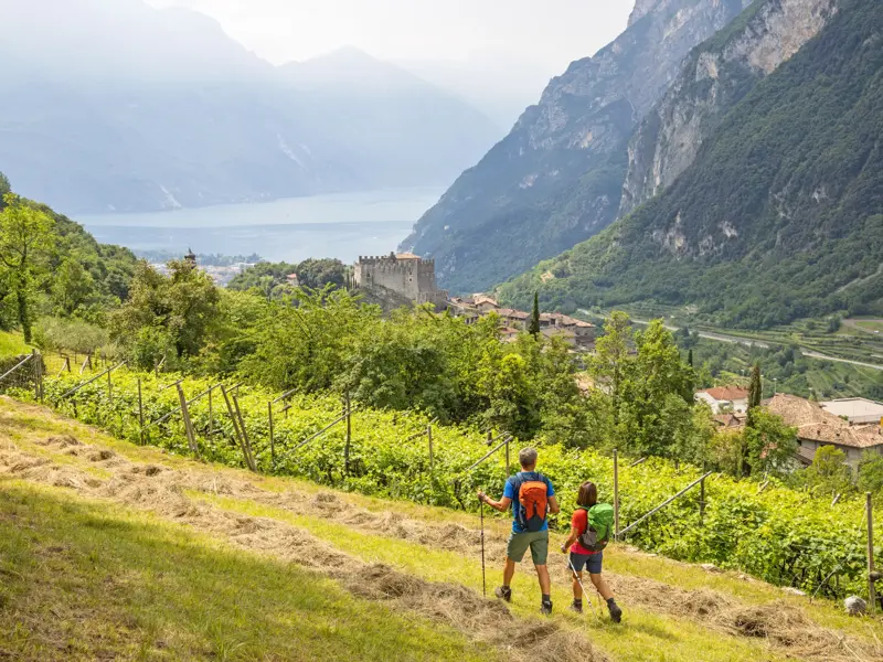Zwei Wanderer mit Rucksäcken blicken beim Abstieg durch Weinberge auf das Castel von Tenno und den Gardasee im Trentino.