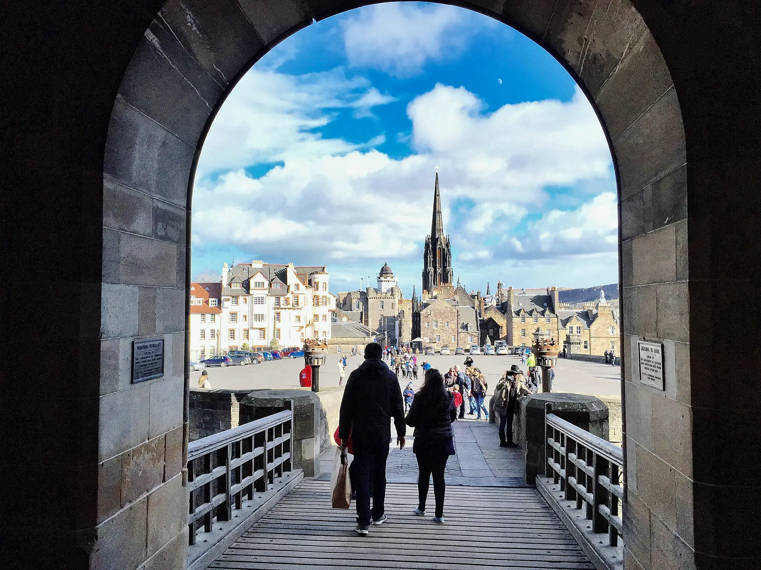Gerahmter Blick durch das Tor des Edinburgh Castle, wo ein Paar auf die belebte Royal Mile mit gotischem Kirchturm blickt.