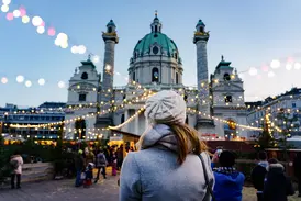 Eine Frau mit Mütze blickt abends über den belebten Weihnachtsmarkt auf die imposante, hell erleuchtete Karlskirche in Wien.