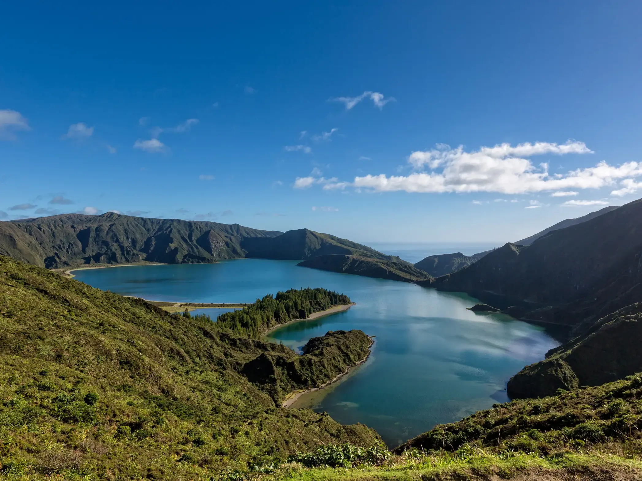Weitblick von einem grünen Berghang auf den tiefblauen Kratersee Lagoa do Fogo auf São Miguel, Azoren.