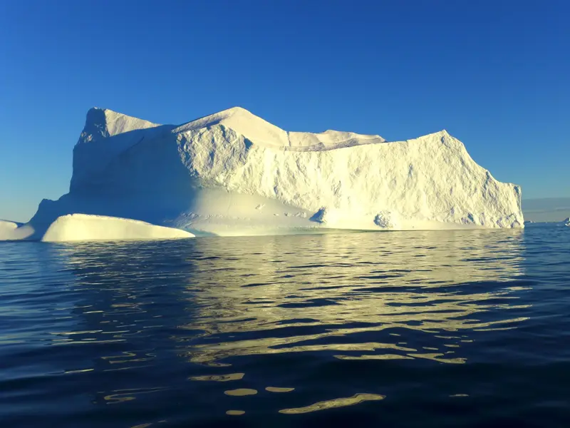 Gewaltiger Eisberg treibt bei Sonnenschein im tiefblauen Wasser eines Fjords während einer Trekkingreise in Ostgrönland.