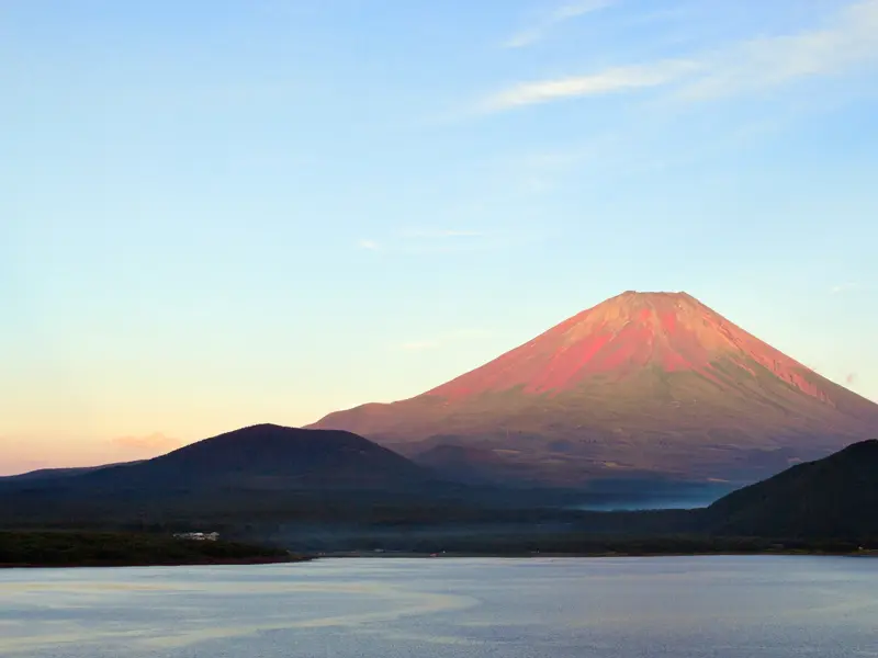 Der Vulkan Fuji-san im Fuji-Hakone-Nationalpark, dessen Gipfel von der Morgensonne rot gefärbt wird, über einem See.