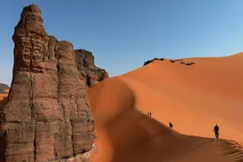 Eine Trekking-Gruppe wandert auf einem hohen Dünenkamm neben bizarren Felsformationen in der Tadrart-Wüste in Algerien.