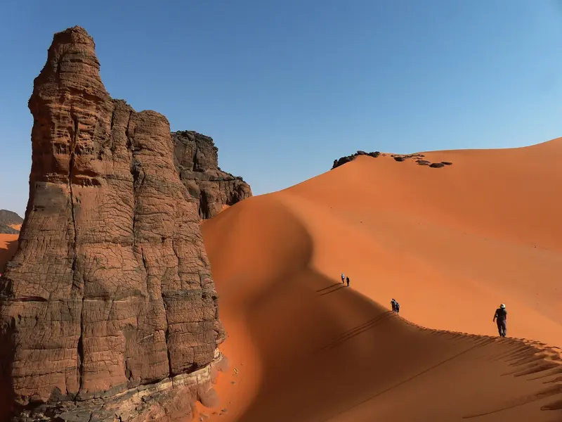 Eine Trekkinggruppe wandert den Grat einer orangefarbenen Sanddüne entlang, vorbei an Felsen im Tassili n'Ajjer Nationalpark.