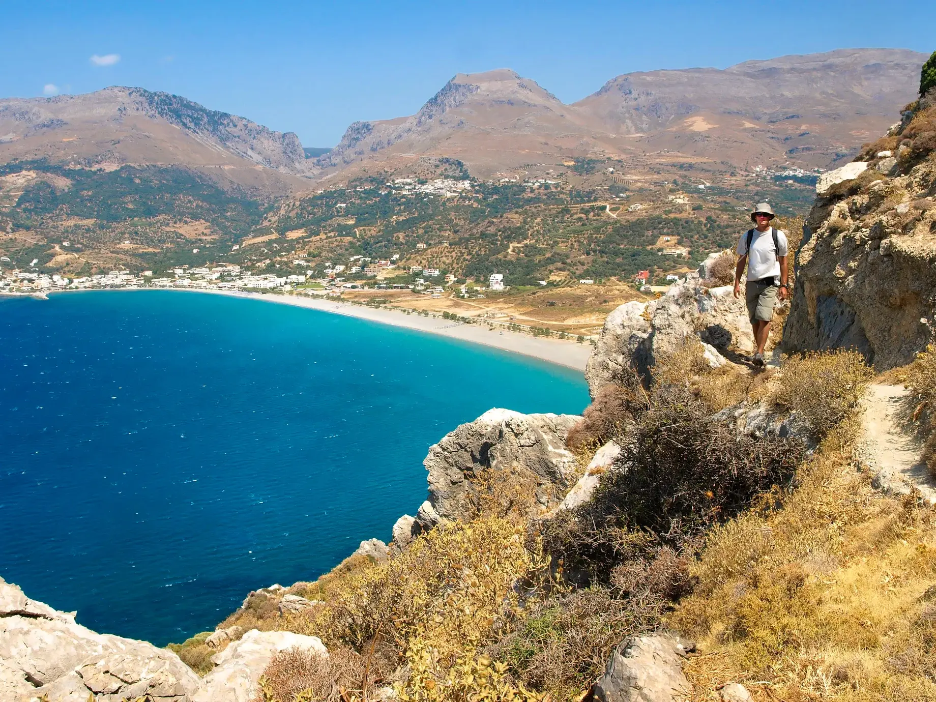 Ein Wanderer mit Rucksack auf einem felsigen Küstenpfad des Fernwanderweges E4 auf Kreta mit Blick auf das türkisblaue Meer.