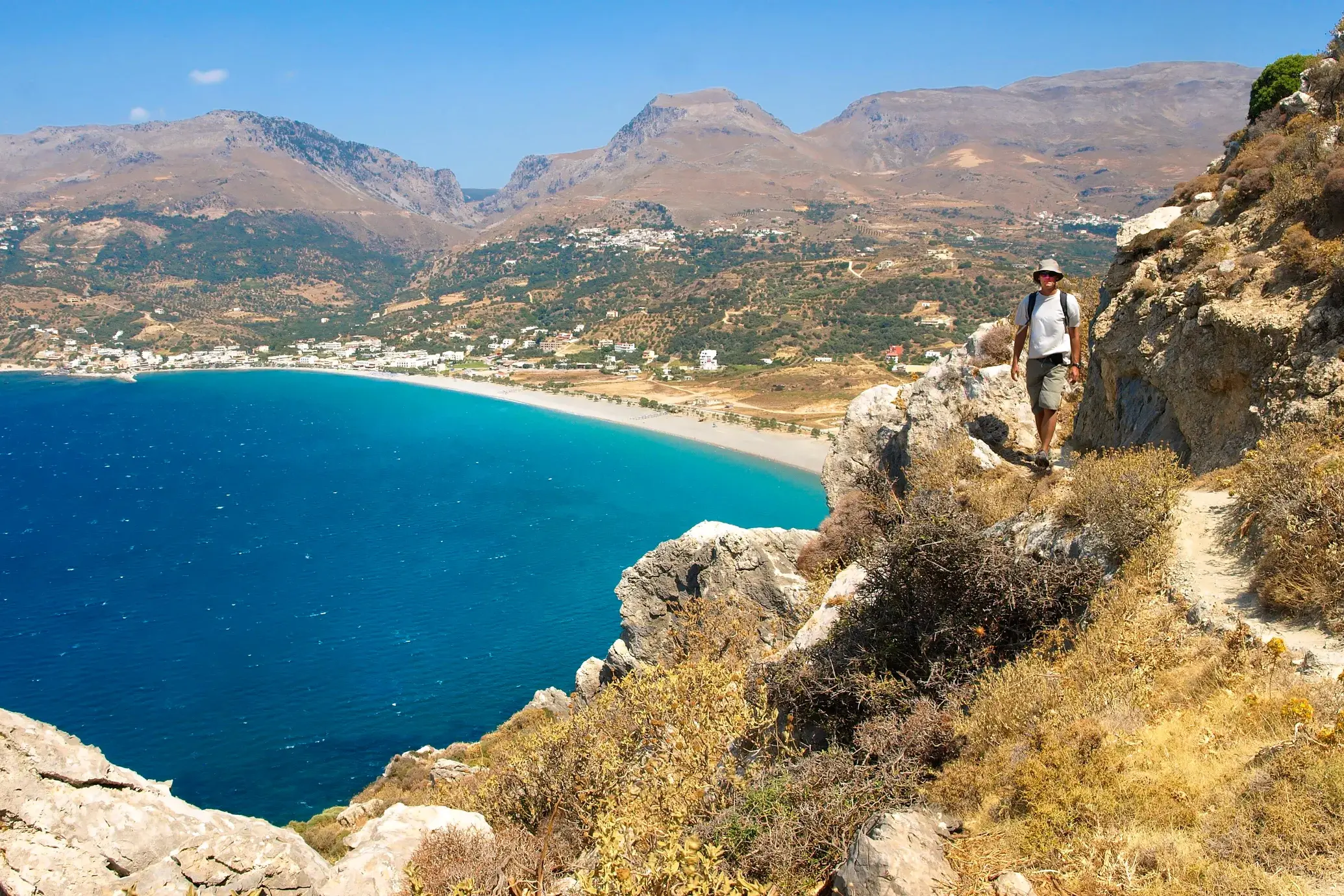 Ein Wanderer mit Rucksack auf einem felsigen Küstenpfad des Fernwanderweges E4 auf Kreta mit Blick auf das türkisblaue Meer.