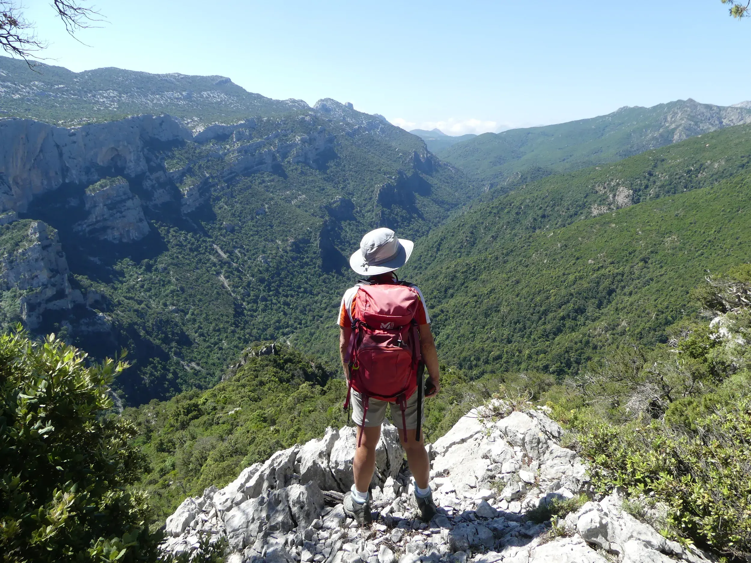 Ein Wanderer mit rotem Rucksack genießt den Ausblick von einem Felsen auf das grüne Tal des Supramonte-Gebirges auf Sardinien.