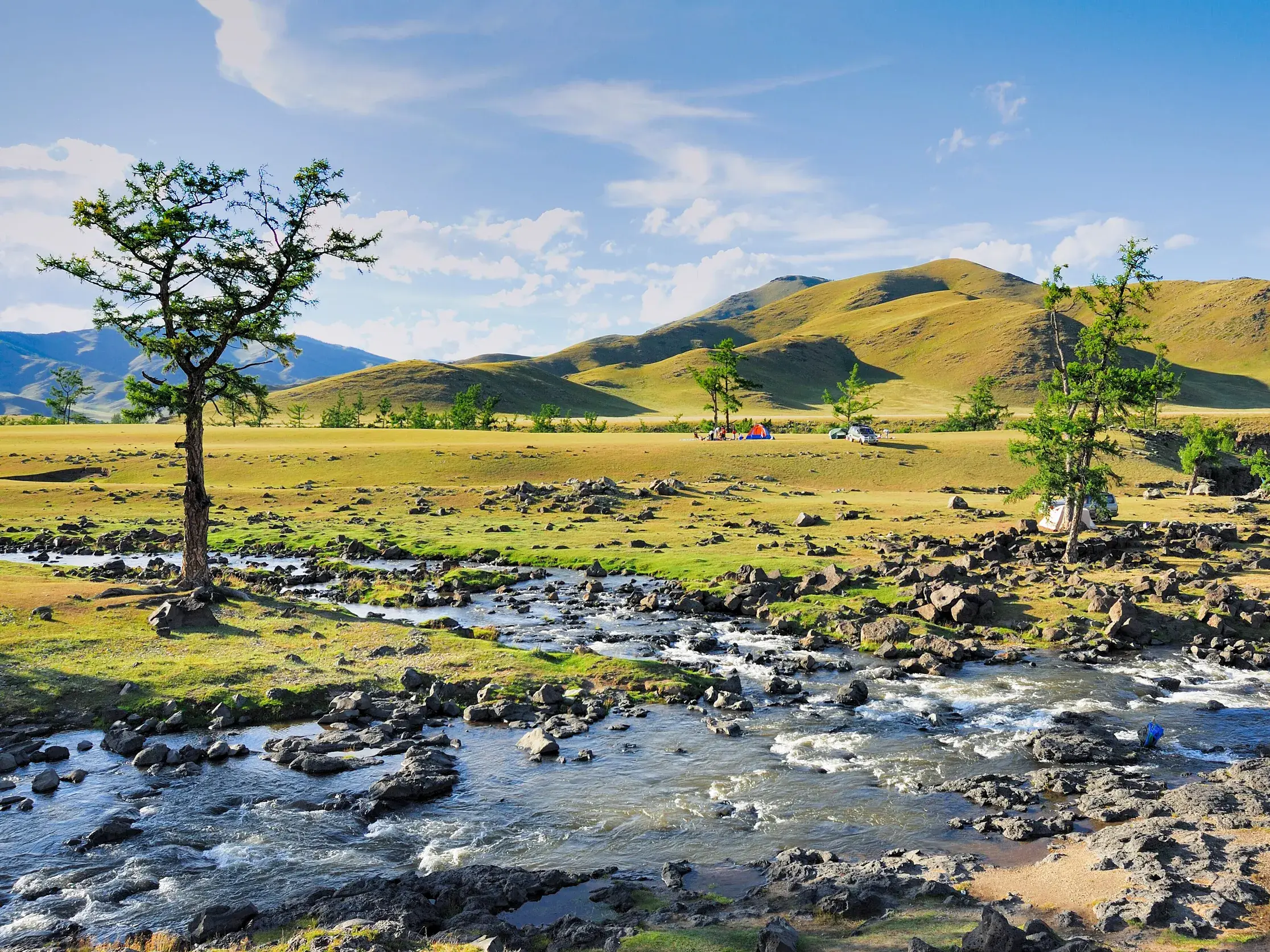 Weite, grüne Steppenlandschaft im Orchon-Tal der Mongolei mit einem klaren Bach und sanften Hügeln am Horizont.