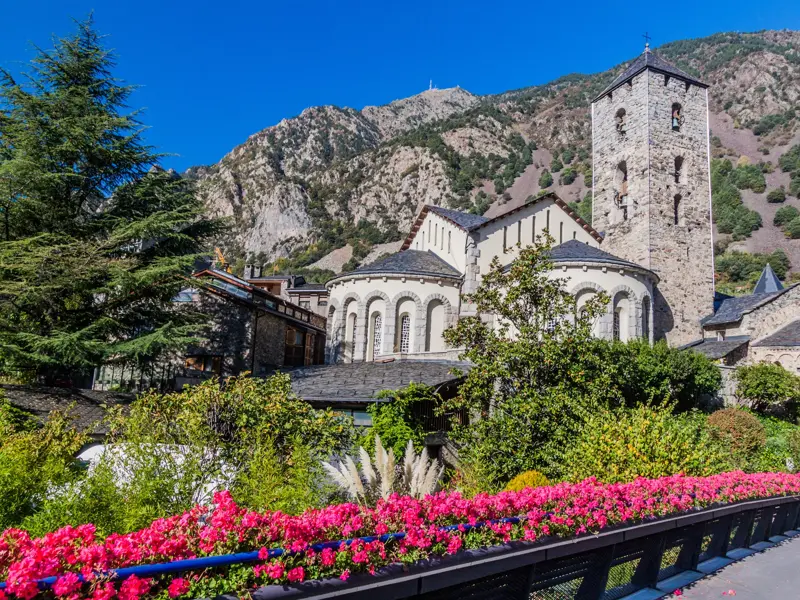 Blick von einer blumengeschmückten Brücke auf die romanische Kirche Santa Coloma in Andorra vor dem Pyrenäen-Bergpanorama.