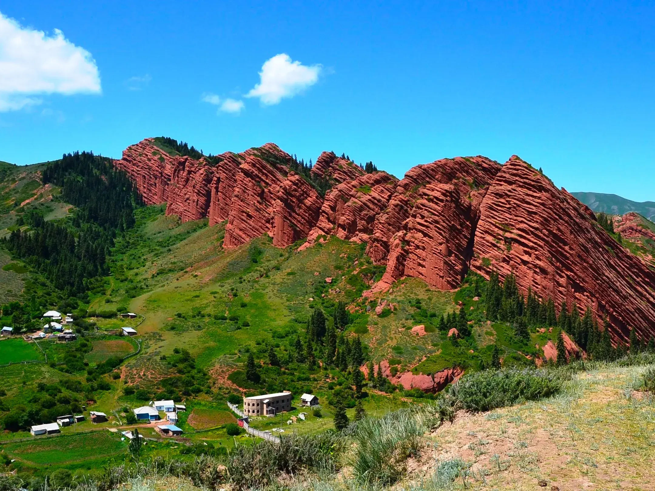 Ein kleines Dorf in einem grünen Tal vor den beeindruckenden roten Felsen des Jeti-Oguz-Tals in Kirgistan.