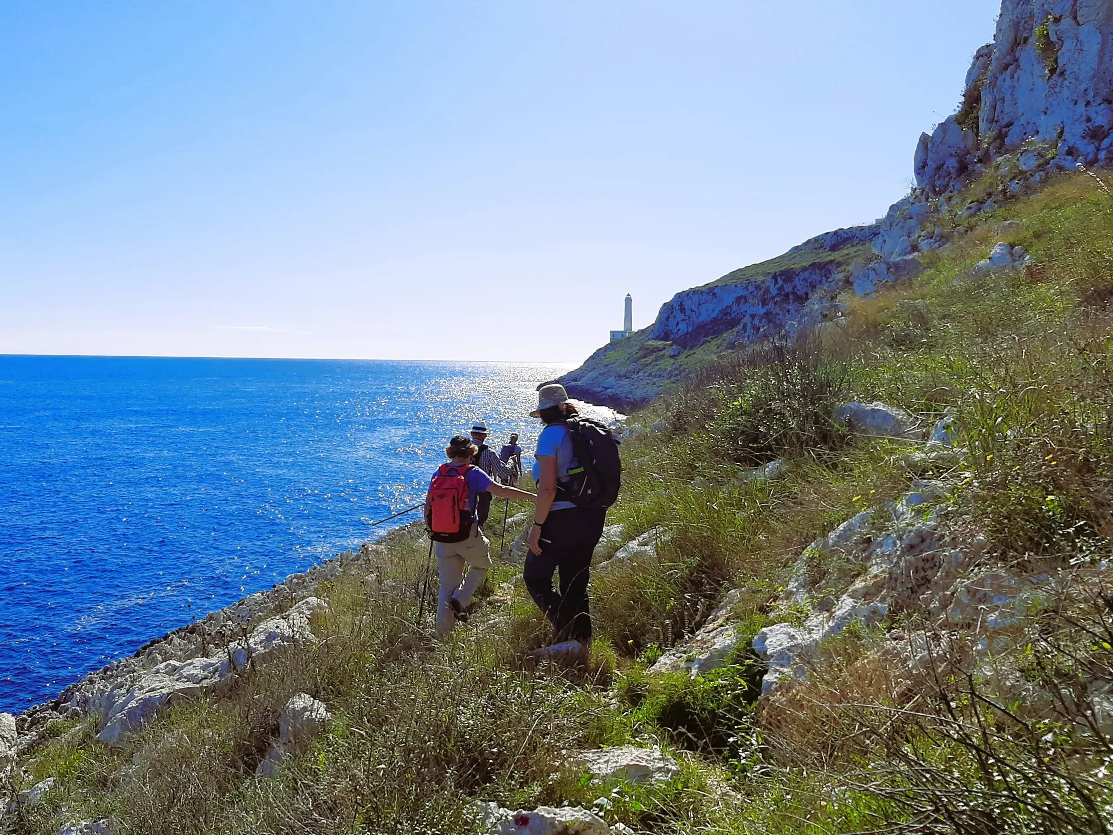 Eine Wandergruppe mit Rucksäcken wandert auf einem Küstenpfad zum Kap von Otranto mit weitem Blick auf das tiefblaue Meer.