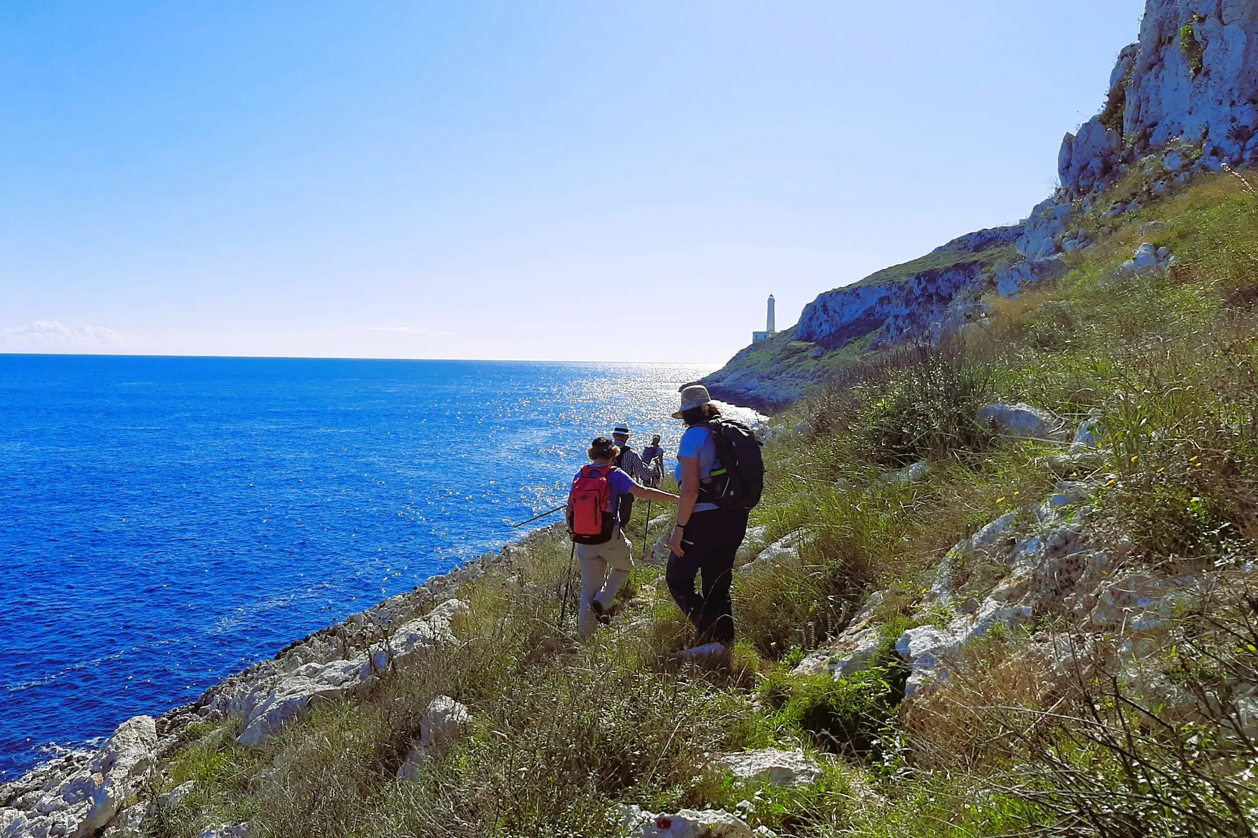 Eine Wandergruppe mit Rucksäcken wandert auf einem Küstenpfad zum Kap von Otranto mit weitem Blick auf das tiefblaue Meer.