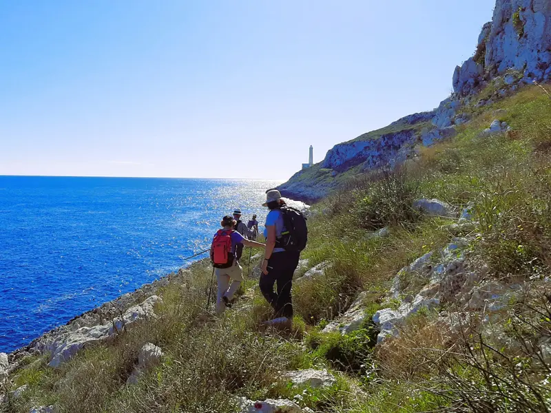Eine Wandergruppe mit Rucksäcken wandert auf einem Küstenpfad zum Kap von Otranto mit weitem Blick auf das tiefblaue Meer.
