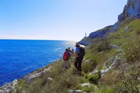 Eine Wandergruppe mit Rucksäcken wandert auf einem Küstenpfad zum Kap von Otranto mit weitem Blick auf das tiefblaue Meer.
