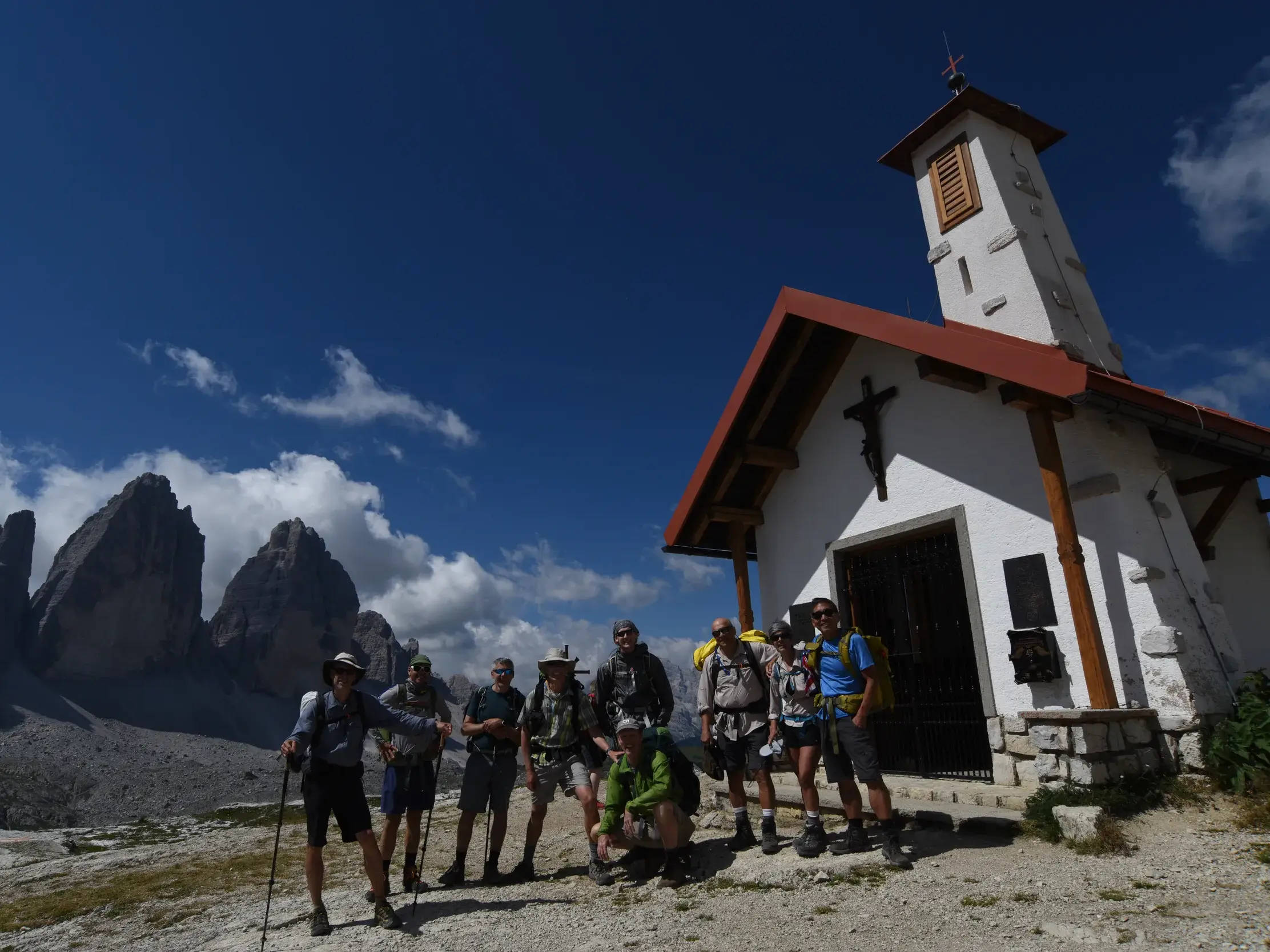 Eine Hauser-Wandergruppe posiert stolz vor einer Bergkapelle mit den berühmten Drei Zinnen im Hintergrund der Dolomiten.