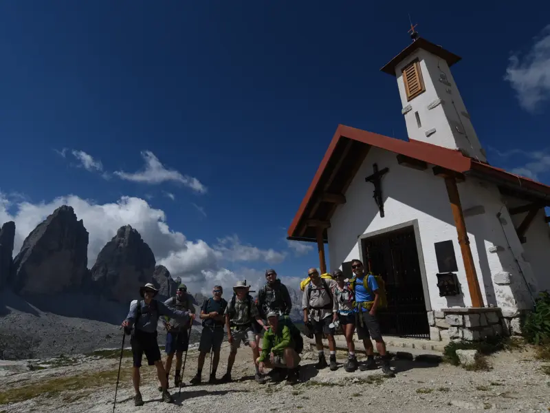 Eine Hauser-Wandergruppe posiert stolz vor einer Bergkapelle mit den berühmten Drei Zinnen im Hintergrund der Dolomiten.