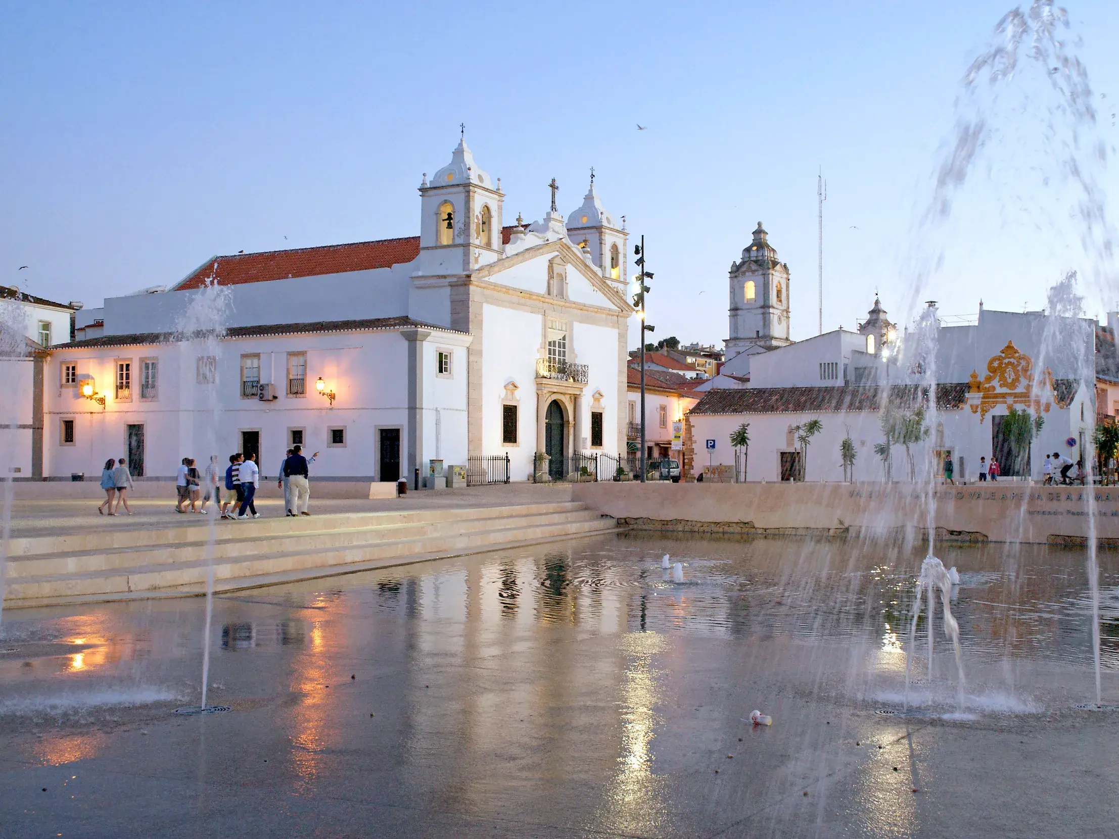 Die weiß getünchte Antoniuskapelle in Lagos an der Algarve bei Dämmerung, davor ein großer Platz mit Wasserspielen.