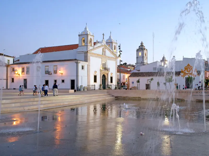 Die weiß getünchte Antoniuskapelle in Lagos an der Algarve bei Dämmerung, davor ein großer Platz mit Wasserspielen.