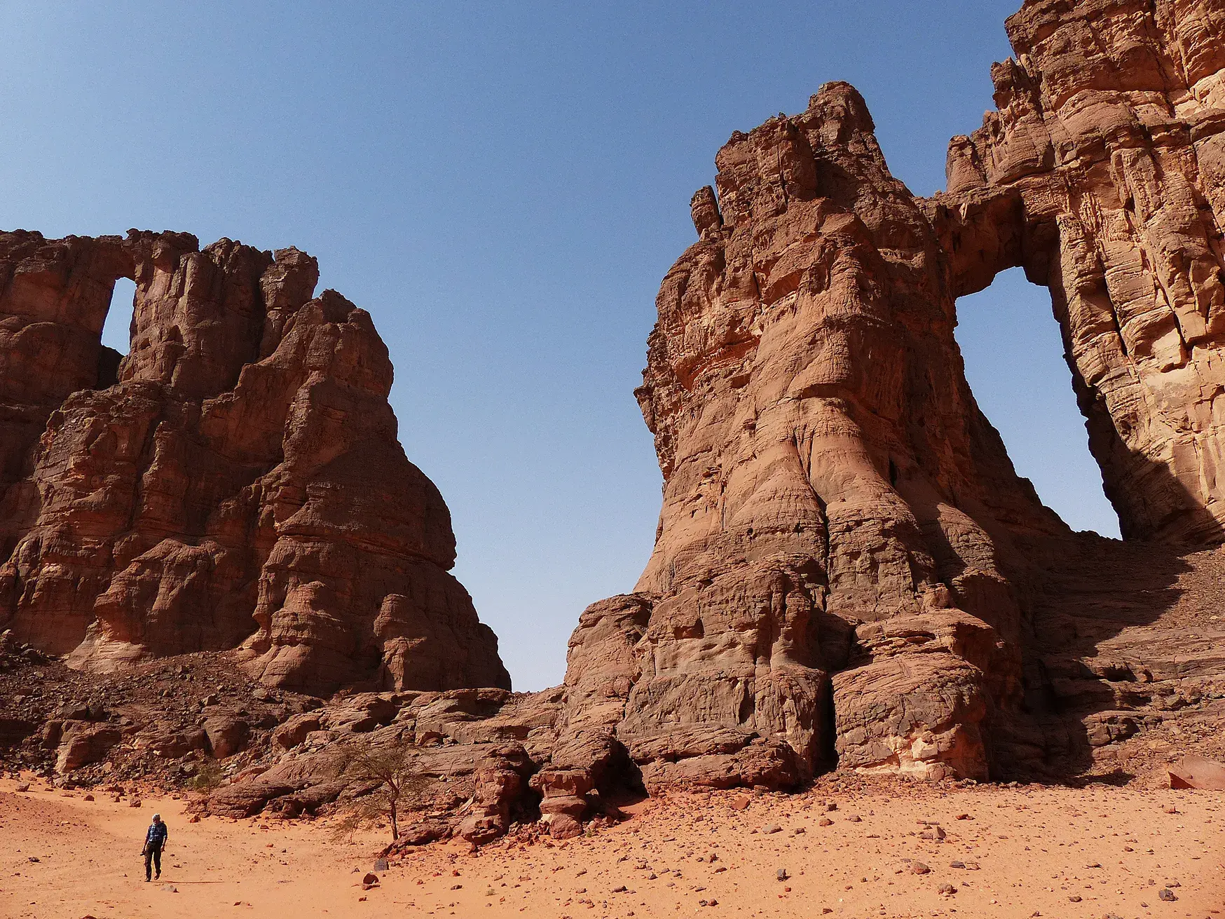 Ein kleiner Wanderer vor gigantischen, roten Felsformationen mit Steinbögen in der Wüstenlandschaft des Tadrart, Algerien.