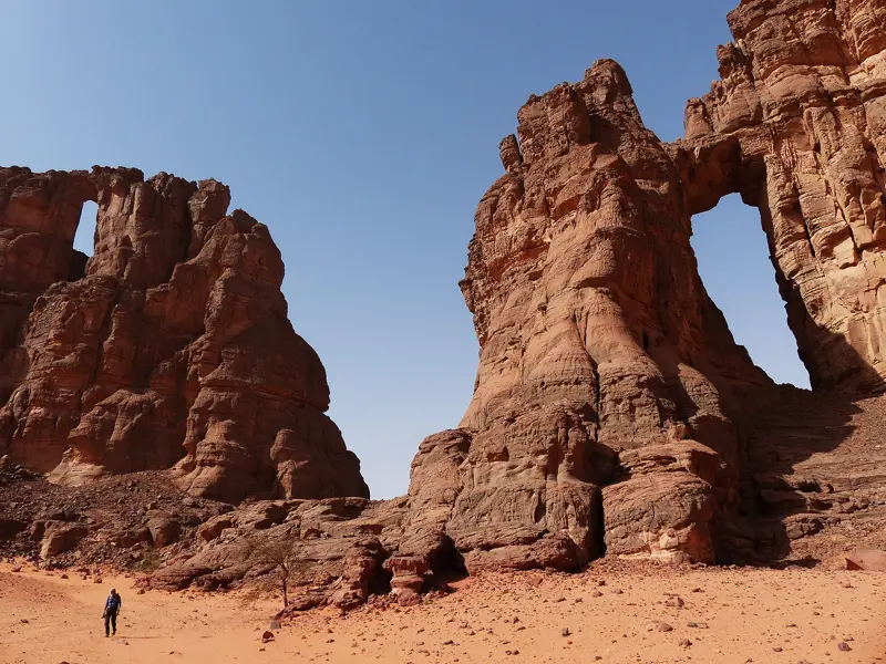 Ein kleiner Wanderer vor gigantischen, roten Felsformationen mit Steinbögen in der Wüstenlandschaft des Tadrart, Algerien.