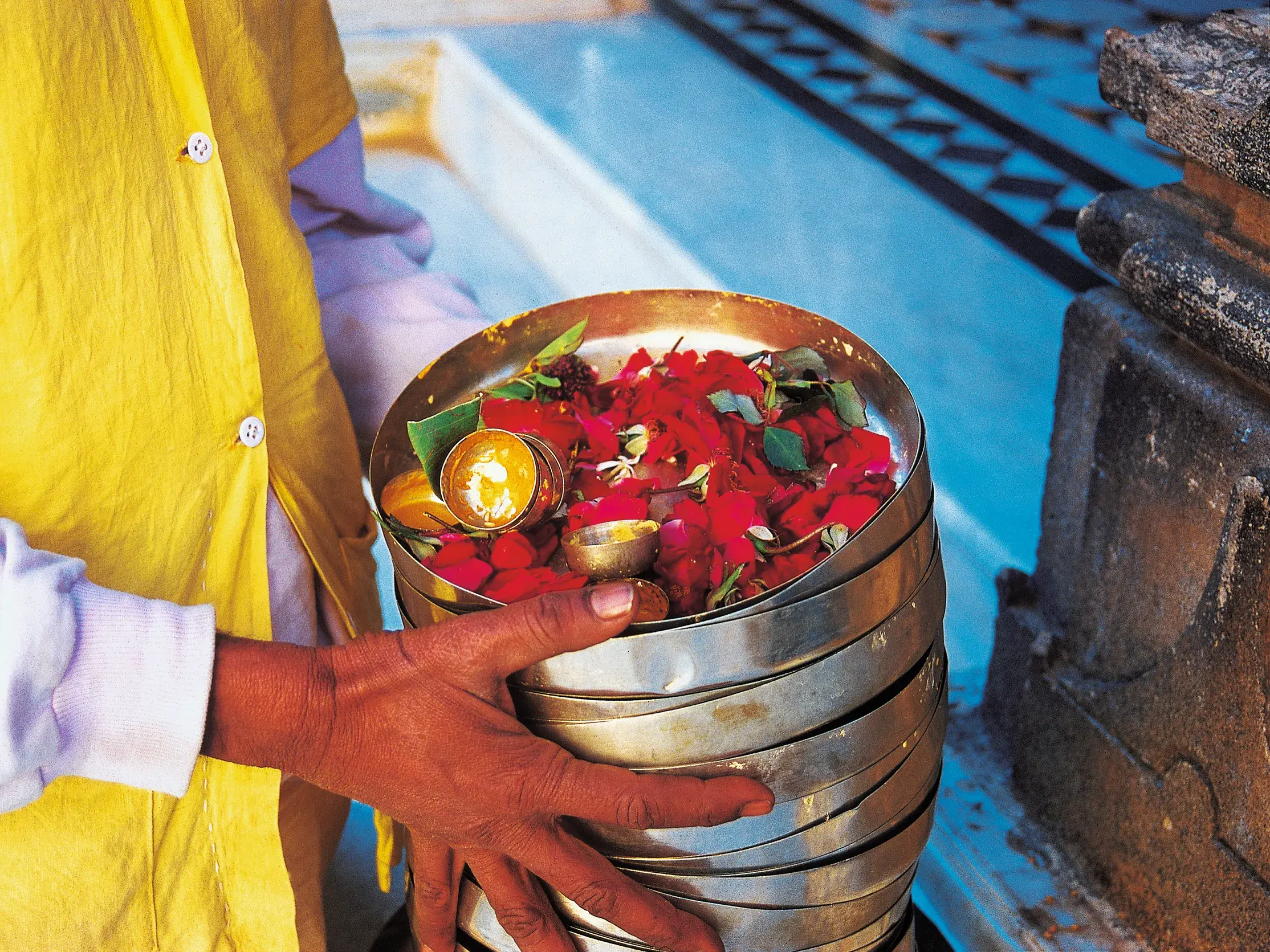 Nahaufnahme einer Hand, die einen Stapel metallener Opferschalen mit roten Blüten für ein spirituelles Ritual in Indien hält.