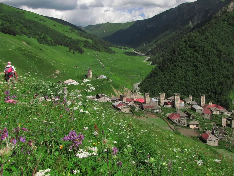Ein Wanderer blickt von einer blühenden Bergwiese auf das von Wehrtürmen geprägte Dorf Uschguli in Swanetien, Georgien.